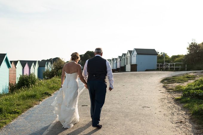 A bride and groom walking hand in hand along a path lined with colourful beach huts in evening light