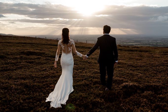 A bride and groom walking hand in hand across moorland toward a dramatic sunset with sunrays breaking through clouds