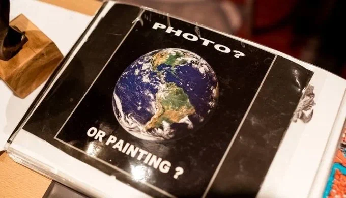 A laminated page in a ring binder showing a photograph of Earth from space, captioned ‘PHOTO?’ above and ‘OR PAINTING?’ below