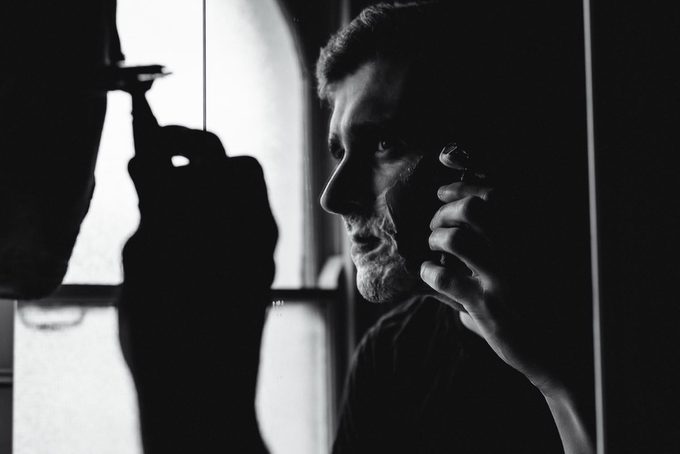 Black and white photo of a man inspecting his shave in a bathroom mirror, with lather remaining on his jaw