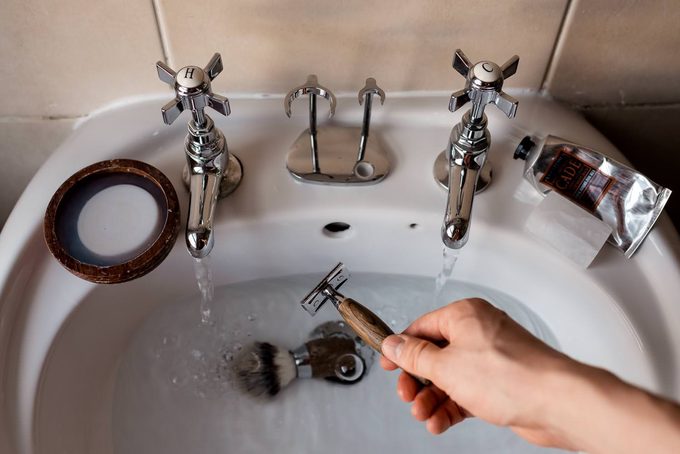 A hand rinsing a wooden-handled safety razor under running water in a bathroom sink, with shaving brush, wooden soap bowl, and aftershave balm nearby